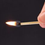 Fingers holding a lit match over black background.